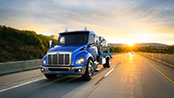 Peterbilt Model 536 Medium Duty Blue Truck with Flatbed Tow / Recovery Body Hauling Car on Highway at Sunset - Thumbnail